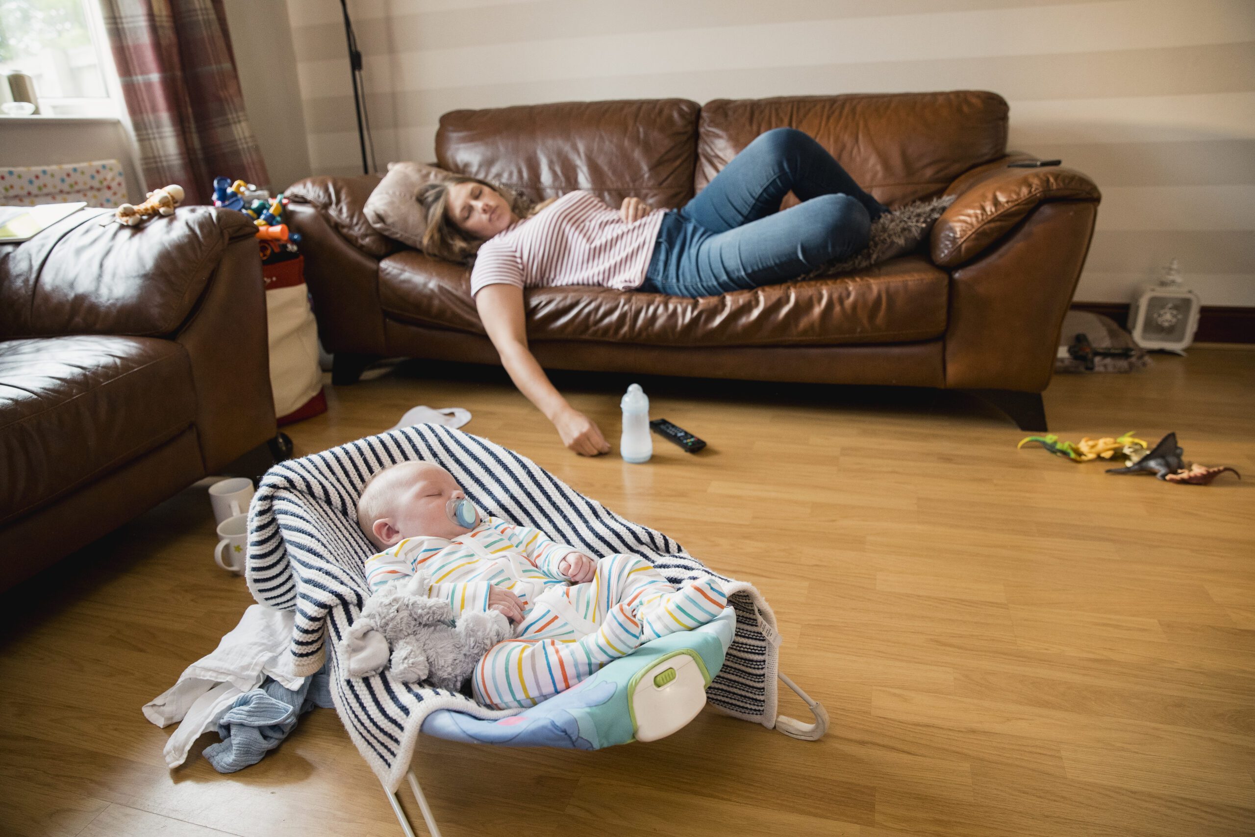 Mother asleep on sofa while baby sleeps nearby in bassinet