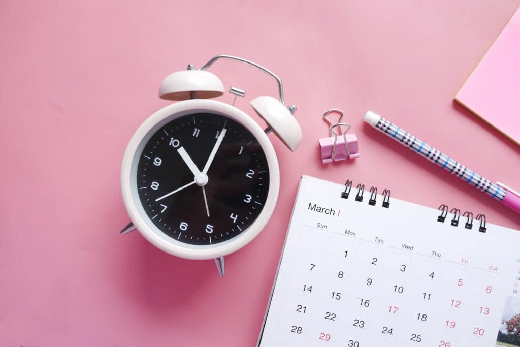 pink analog alarm clock, flipbook calendar, plaid pen, and pink binder clip on pink background