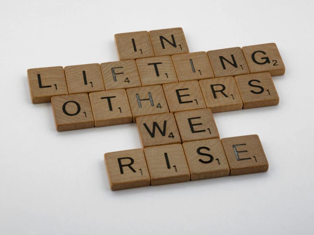 Brown wooden scrabble tiles reading "in lifting others we rise".