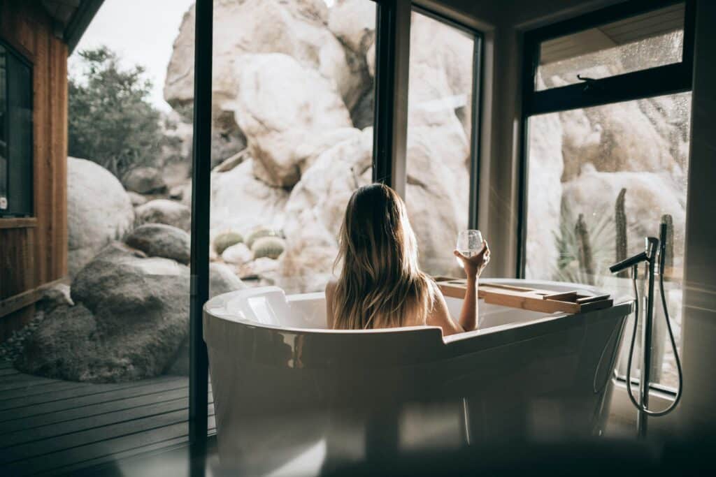 Woman in white bathtub holding wine glass looking out at desert landscape.